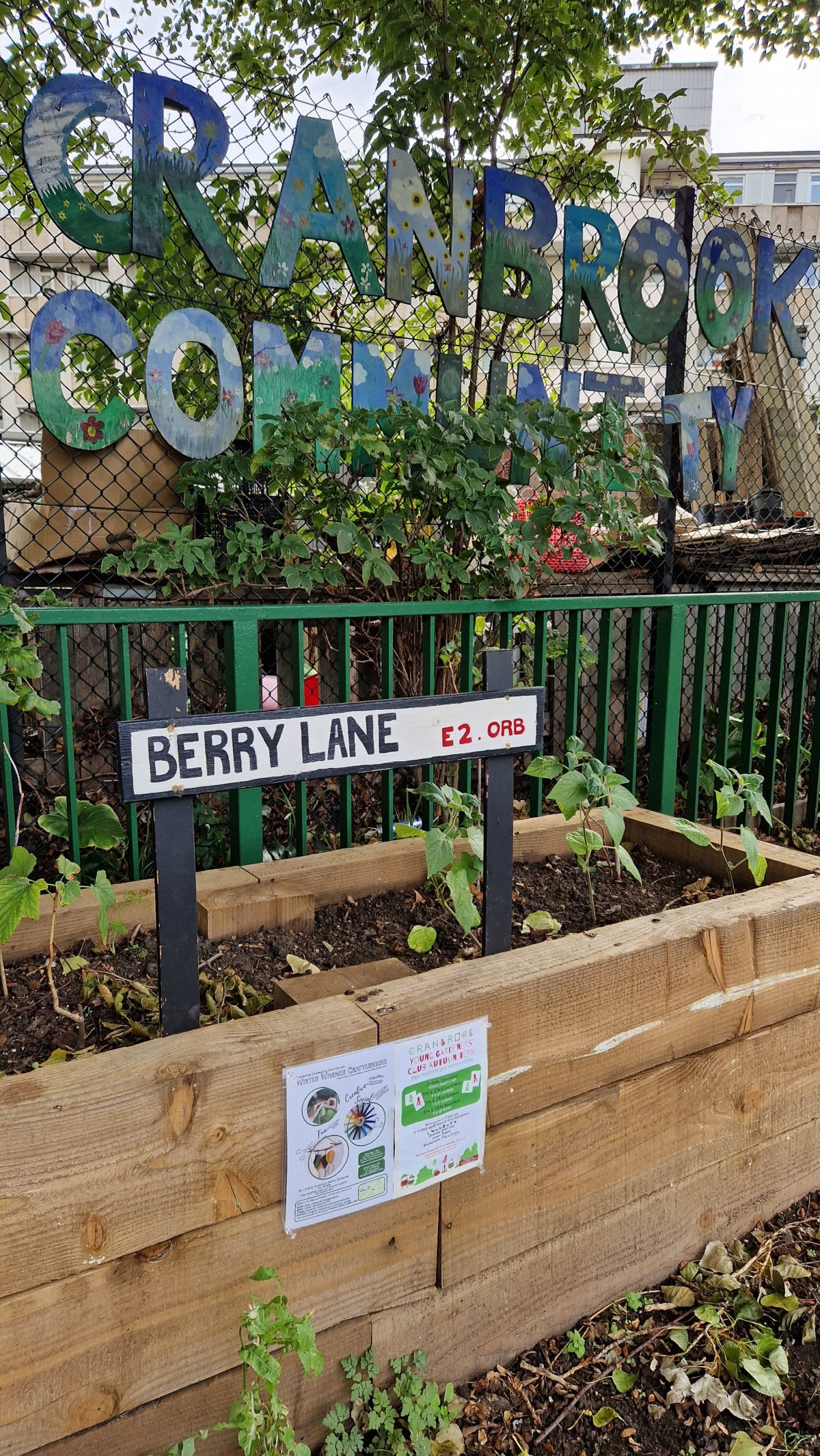 A large planter made of thick pieces of wood contains a sign painted to look like a street sign. It says "Berry Lane". Behind it is a green metal fence.
