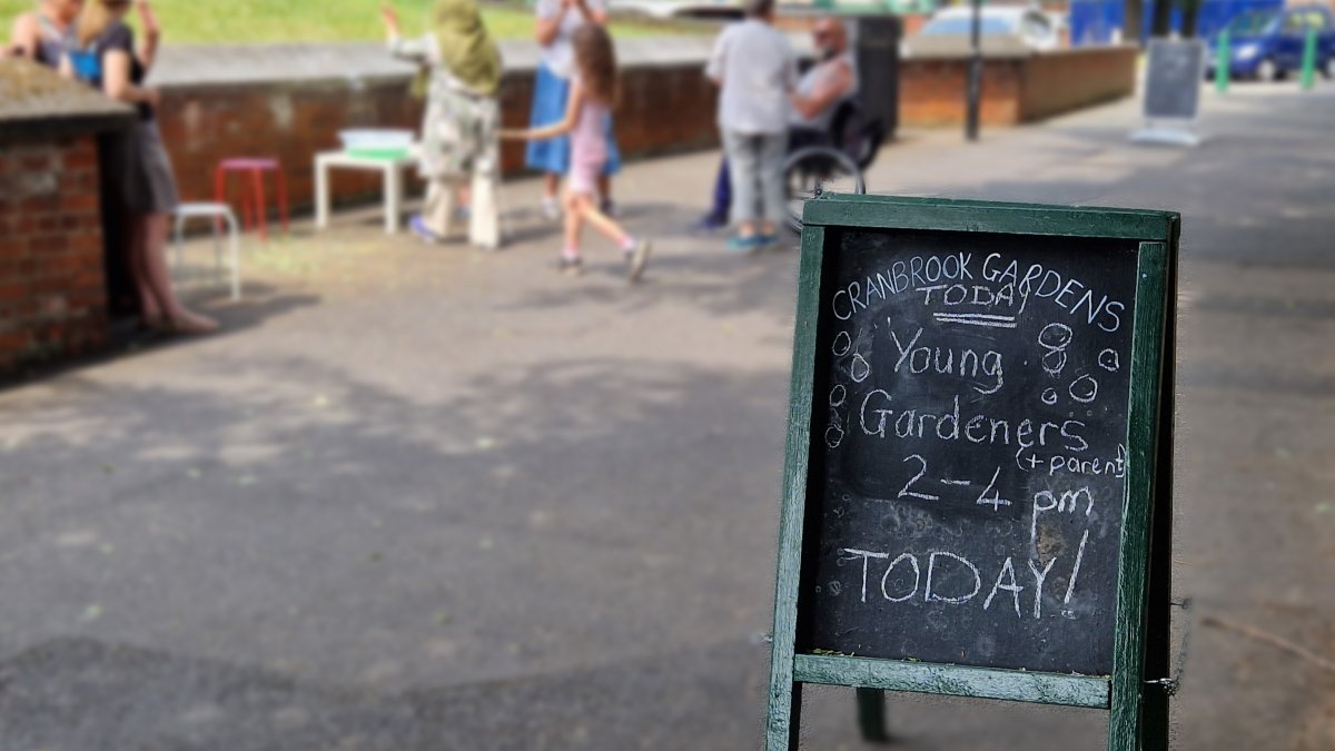 A sign saying "Young Gardeners 2-4pm today" stands at the side of an open footpath. In the background people are chatting while children blow bubbles.