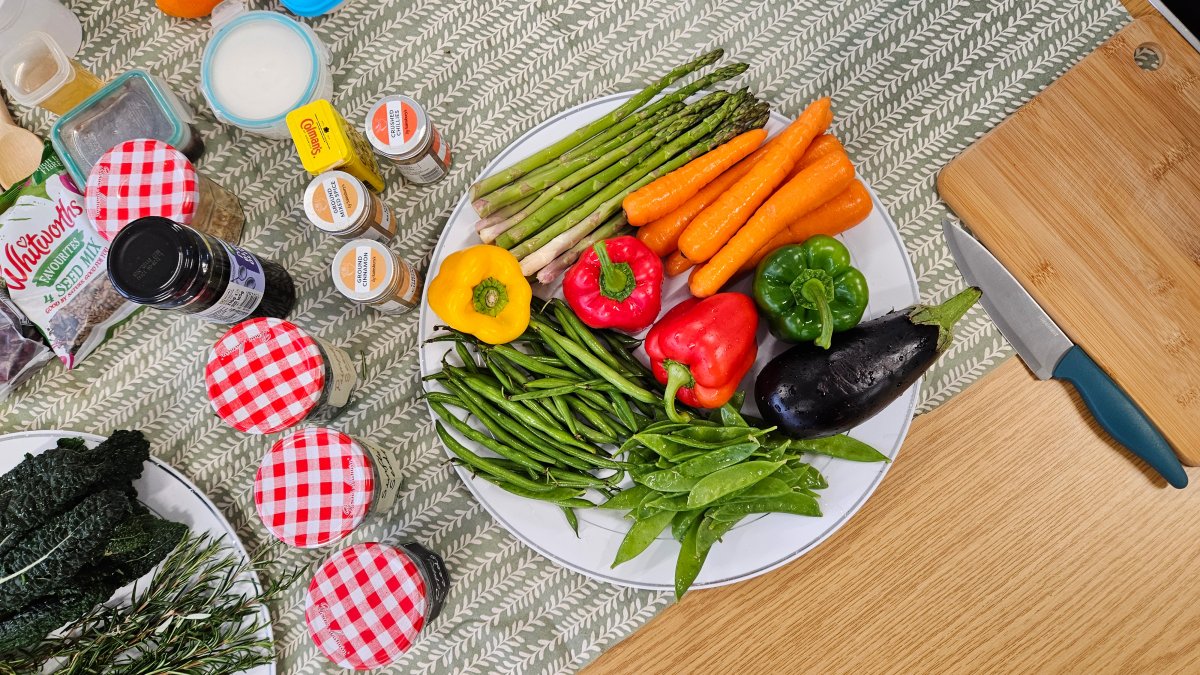 A selection of bright colourful fresh vegetables sits on a plate surrounded by jars of seeds and spices.
